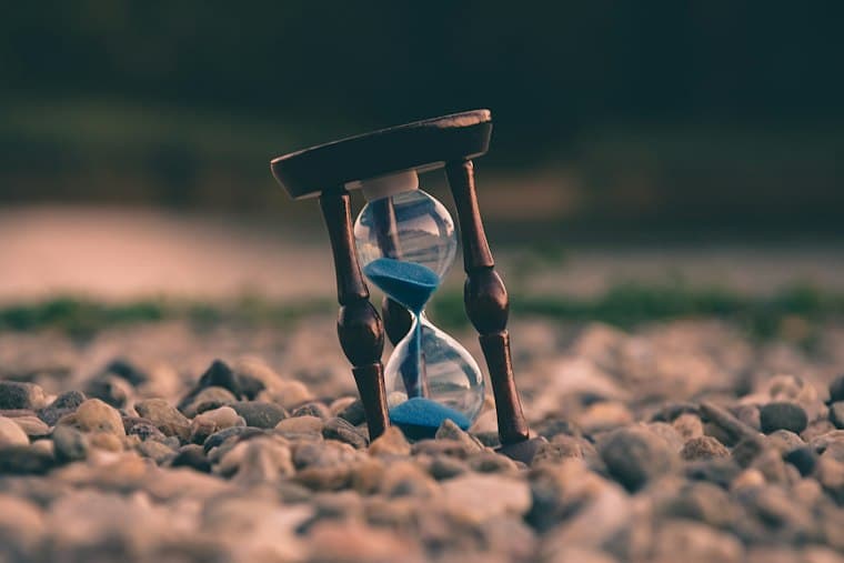 A wooden hourglass with blue sand standing on a bed of pebbles