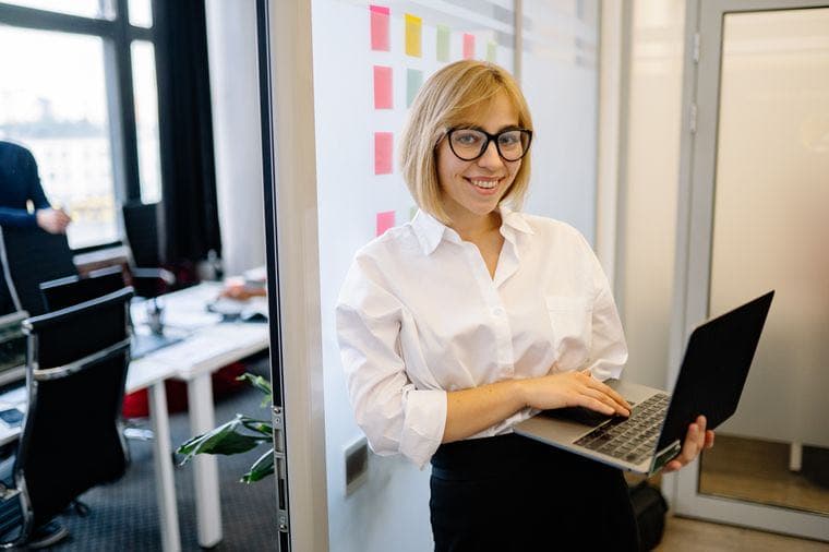 A confident smiling woman holding a laptop in an office