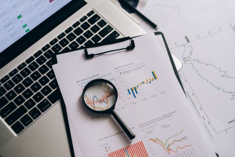 A magnifying glass held over printed financial charts beside a laptop