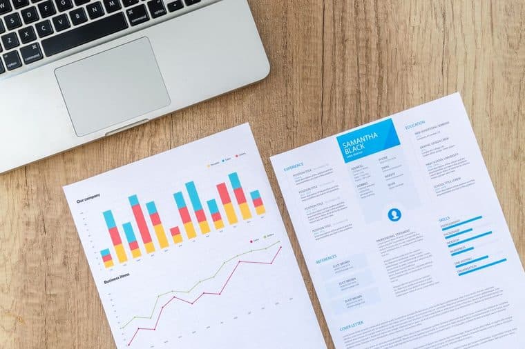 A resume and charts on a desk next to a laptop during a job search