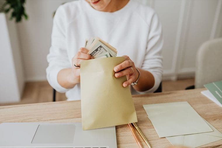 A person sliding banknotes into an envelope at a desk, more envelopes waiting beside it