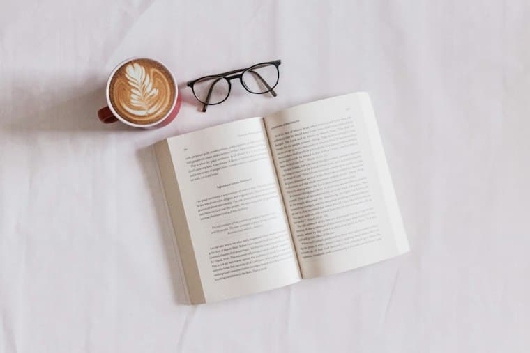 An open book beside a latte and reading glasses on a white sheet
