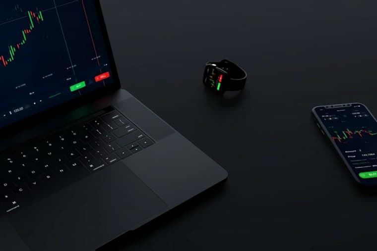 A laptop, watch and phone on a dark desk, all showing trading charts
